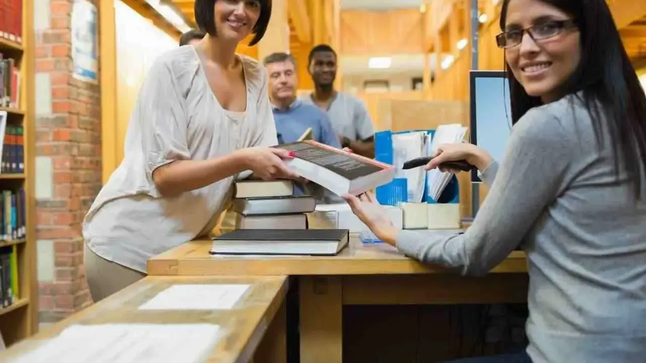 Due donne sorridono mentre lavorano in biblioteca, una passa un libro, l'altra usa uno scanner.