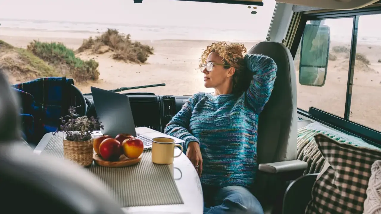 Donna con capelli ricci lavora al computer in un camper, godendosi la vista del mare. Un esempio di nomade digitale lavoro.