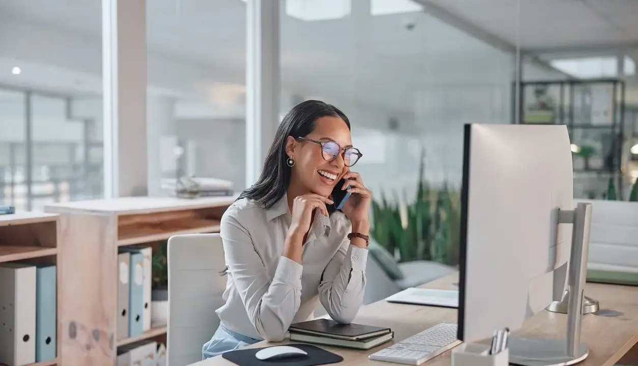 Sorride al telefono, una segretaria part time mattina, mentre lavora al computer in un ufficio moderno.