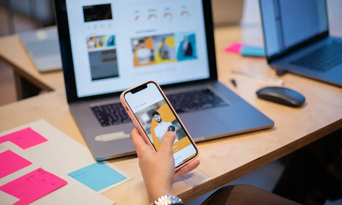 Un ux designer valuta un'interfaccia utente su smartphone, con laptop e post-it colorati sulla scrivania.
