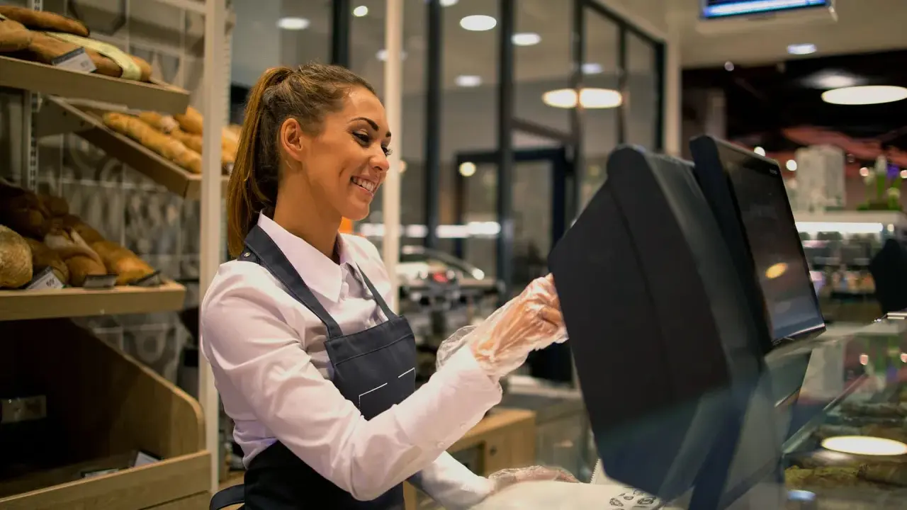 Ragazza sorridente con grembiule, guanti e camicia bianca, pronta per il suo corso gratuito addetto alla cassa in un panificio.