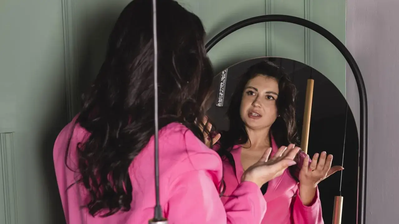 Donna in giacca rosa si guarda allo specchio, sembra parlare da sola e gesticolare con le mani, come se stesse provando un discorso.