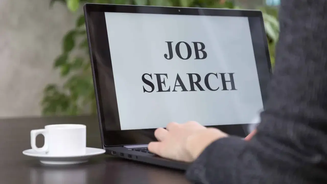 Persona che digita su un laptop con scritto "JOB SEARCH". Un caffè è accanto, pronto per iniziare a cercare lavoro.