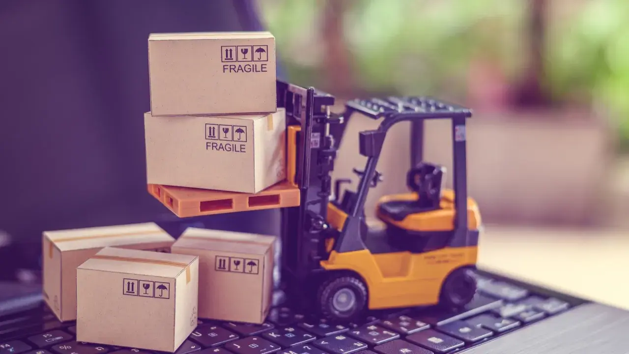 Carrellino elevatore giallo solleva scatole fragili su un pallet, mentre altre scatole attendono su una tastiera. Il buyer cosa fa? Organizza la spedizione.