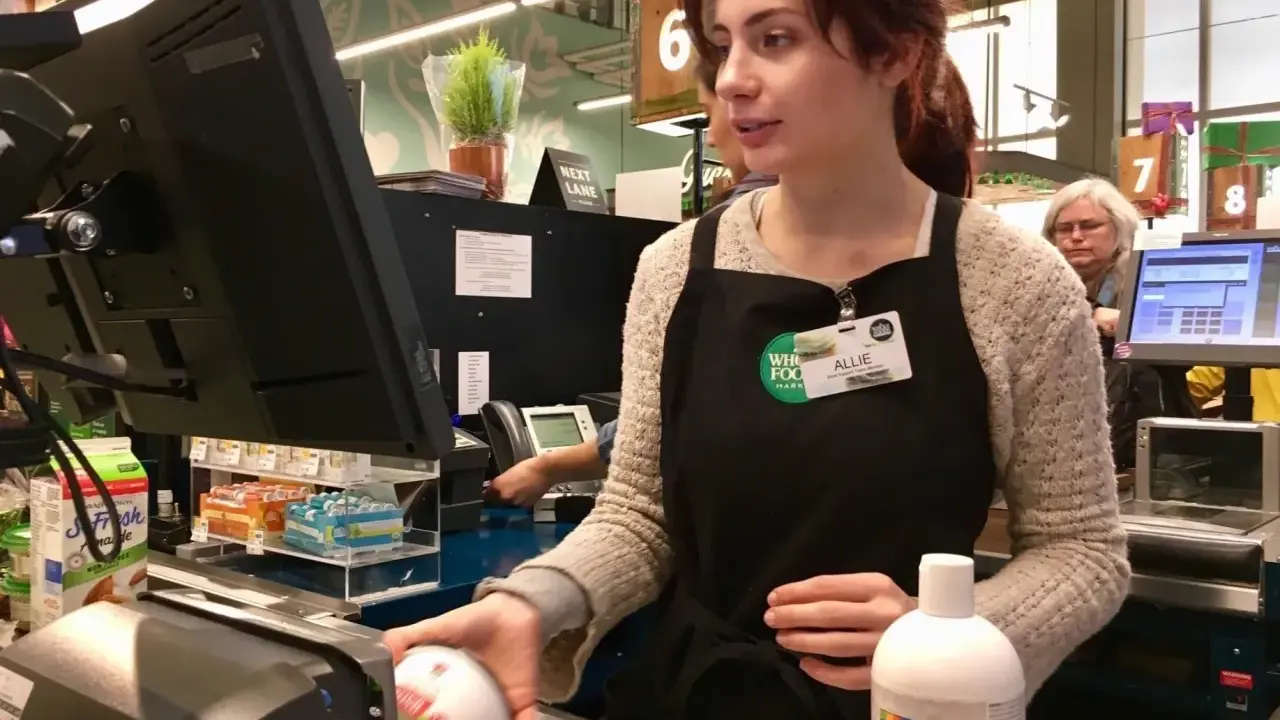 Cassiera al supermercato, Allie, con il suo nome visibile, lavora al registratore di cassa. Il suo stipendio da cassiera di supermercato è guadagnato con ogni transazione.