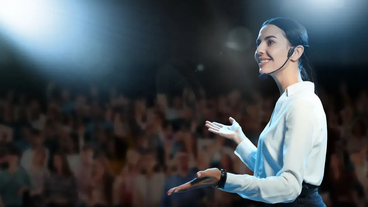 Oratrice sorridente sul palco, con microfono, che parla a un pubblico. Nonostante l'ansia, non riesco a parlare in modo fluido, ma cerco di comunicare.