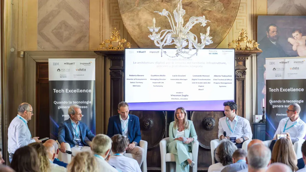 Panel di esperti discute di architetture digitali. Uno dei relatori, con un completo blu, sembra pensare: "non so che lavoro fare".