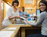 Due donne sorridono mentre lavorano in biblioteca, una passa un libro, l'altra usa uno scanner.