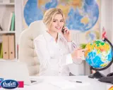 Donna sorridente al telefono, indica un globo terrestre. L'ufficio è arredato per un corso agenzia viaggi.