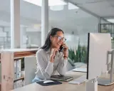Sorride al telefono, una segretaria part time mattina, mentre lavora al computer in un ufficio moderno.