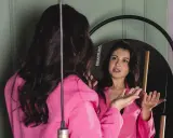 Donna in giacca rosa si guarda allo specchio, sembra parlare da sola e gesticolare con le mani, come se stesse provando un discorso.