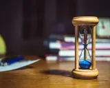 Clessidra con sabbia blu su una scrivania, a simboleggiare quante ore devono passare tra un turno e l'altro. Libri e fogli sullo sfondo.