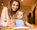 Un educatore professionale guida una bambina nel disegno, con matite colorate sparse sul tavolo.