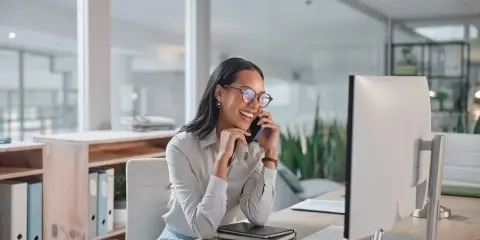 Sorride al telefono, una segretaria part time mattina, mentre lavora al computer in un ufficio moderno.