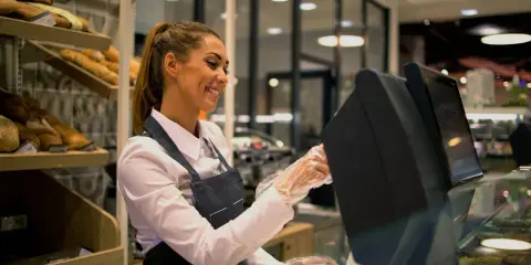 Ragazza sorridente con grembiule, guanti e camicia bianca, pronta per il suo corso gratuito addetto alla cassa in un panificio.
