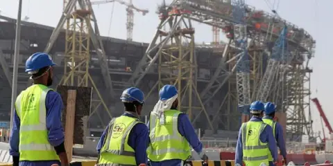 Operai con caschi e giubbotti ad alta visibilità lavorano in Qatar alla costruzione di un imponente stadio.