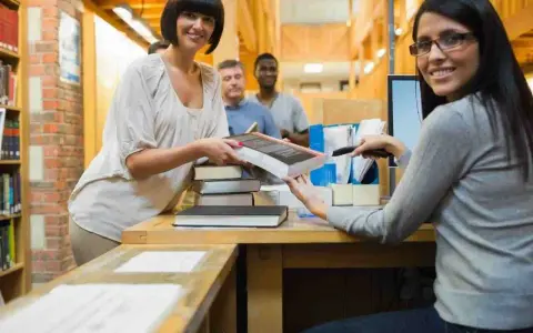Due donne sorridono mentre lavorano in biblioteca, una passa un libro, l'altra usa uno scanner.