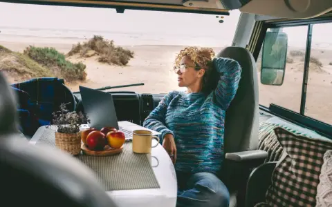 Donna con capelli ricci lavora al computer in un camper, godendosi la vista del mare. Un esempio di nomade digitale lavoro.