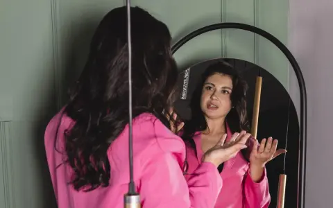 Donna in giacca rosa si guarda allo specchio, sembra parlare da sola e gesticolare con le mani, come se stesse provando un discorso.