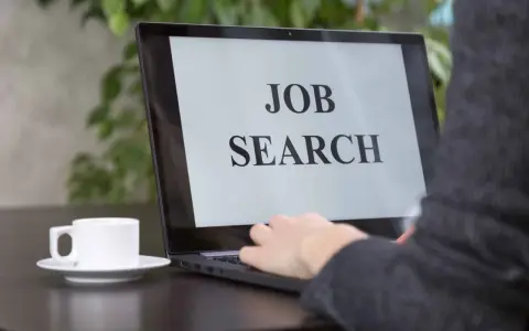 Persona che digita su un laptop con scritto "JOB SEARCH". Un caffè è accanto, pronto per iniziare a cercare lavoro.