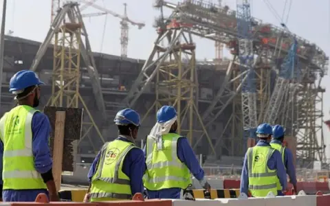 Operai con caschi e giubbotti ad alta visibilità lavorano in Qatar alla costruzione di un imponente stadio.