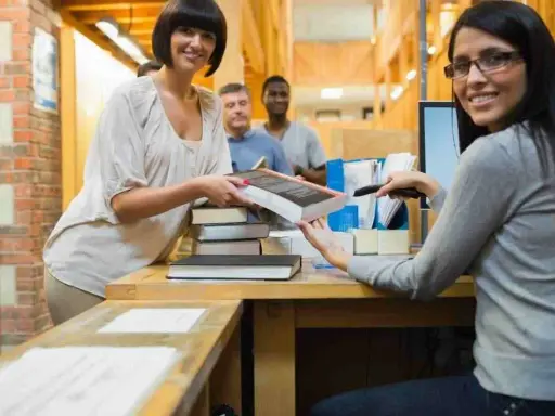 Due donne sorridono mentre lavorano in biblioteca, una passa un libro, l'altra usa uno scanner.