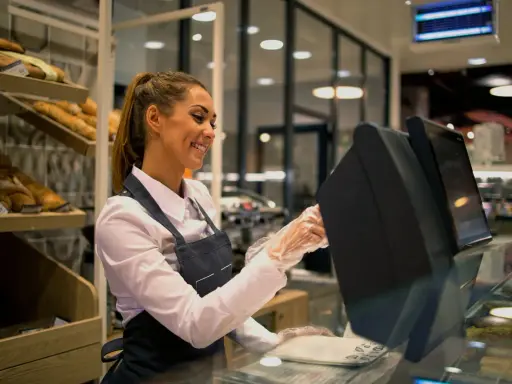 Ragazza sorridente con grembiule, guanti e camicia bianca, pronta per il suo corso gratuito addetto alla cassa in un panificio.