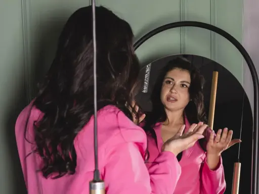 Donna in giacca rosa si guarda allo specchio, sembra parlare da sola e gesticolare con le mani, come se stesse provando un discorso.