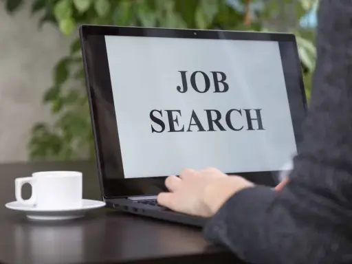 Persona che digita su un laptop con scritto "JOB SEARCH". Un caffè è accanto, pronto per iniziare a cercare lavoro.