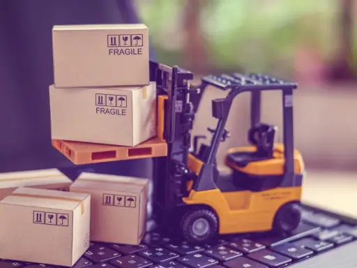 Carrellino elevatore giallo solleva scatole fragili su un pallet, mentre altre scatole attendono su una tastiera. Il buyer cosa fa? Organizza la spedizione.