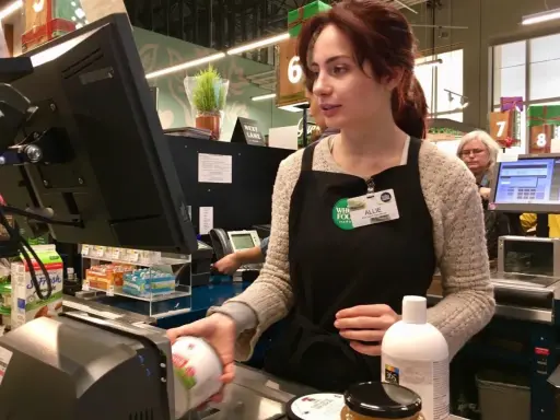 Cassiera al supermercato, Allie, con il suo nome visibile, lavora al registratore di cassa. Il suo stipendio da cassiera di supermercato è guadagnato con ogni transazione.