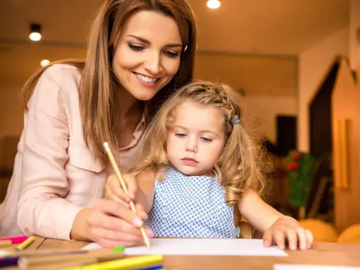 Un educatore professionale guida una bambina nel disegno, con matite colorate sparse sul tavolo.