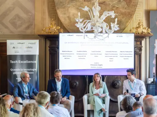 Panel di esperti discute di architetture digitali. Uno dei relatori, con un completo blu, sembra pensare: "non so che lavoro fare".