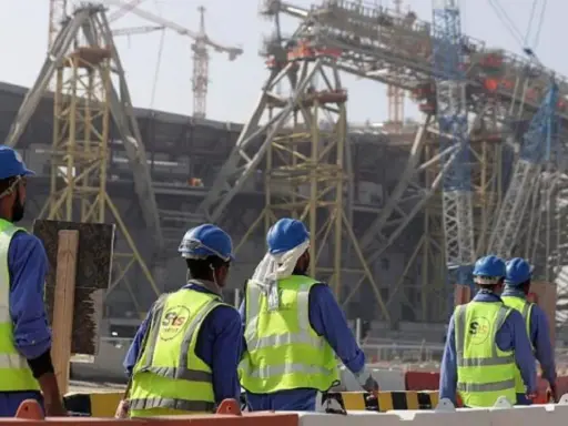 Operai con caschi e giubbotti ad alta visibilità lavorano in Qatar alla costruzione di un imponente stadio.