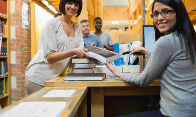 Due donne sorridono mentre lavorano in biblioteca, una passa un libro, l'altra usa uno scanner.