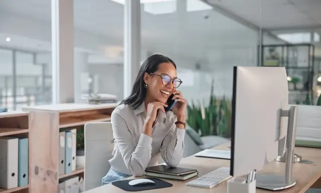 Sorride al telefono, una segretaria part time mattina, mentre lavora al computer in un ufficio moderno.