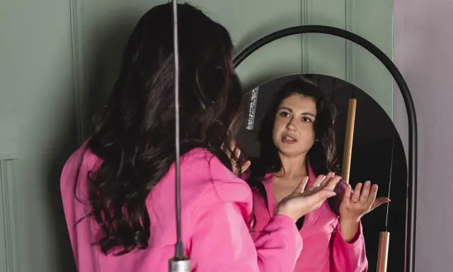 Donna in giacca rosa si guarda allo specchio, sembra parlare da sola e gesticolare con le mani, come se stesse provando un discorso.