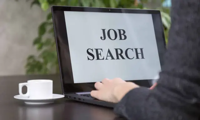 Persona che digita su un laptop con scritto "JOB SEARCH". Un caffè è accanto, pronto per iniziare a cercare lavoro.