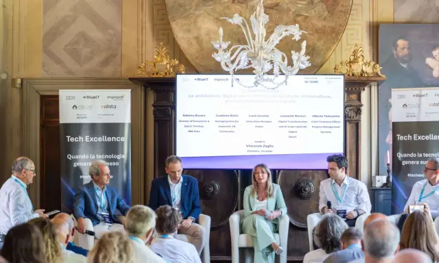Panel di esperti discute di architetture digitali. Uno dei relatori, con un completo blu, sembra pensare: "non so che lavoro fare".