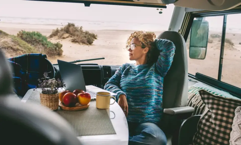 Donna con capelli ricci lavora al computer in un camper, godendosi la vista del mare. Un esempio di nomade digitale lavoro.