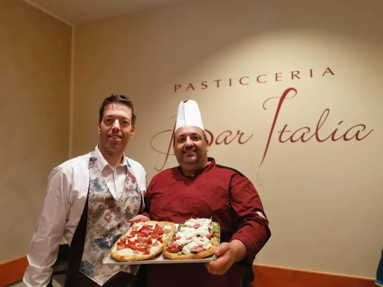 Un cuoco e un banconista sorridono mostrando due focacce. Il banconista cosa fa? Prepara e serve delizie!