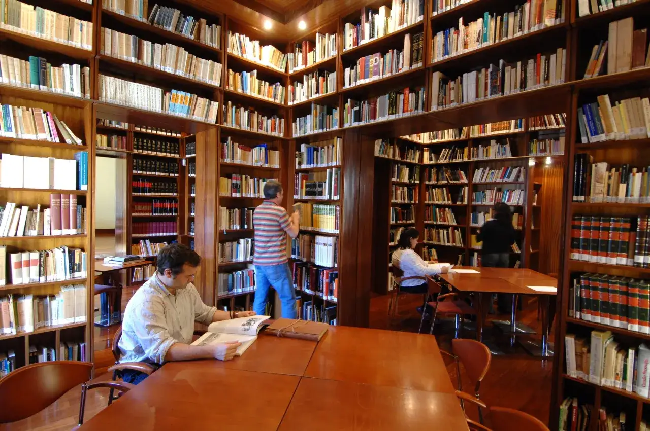 Persone che studiano e lavorano in biblioteca, circondate da scaffali pieni di libri.