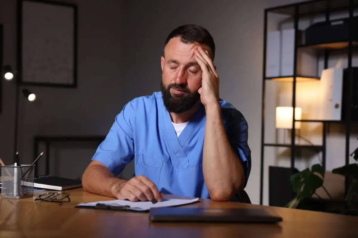 Medico esausto, con la mano sulla fronte, mentre lavora di notte. La stanchezza è palpabile mentre esamina documenti alla scrivania.