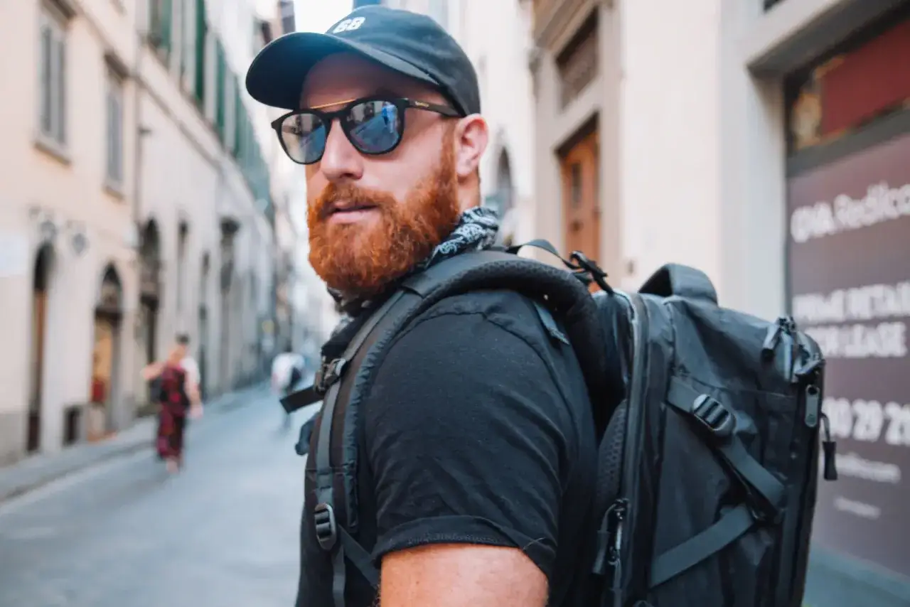 Uomo con barba rossa, occhiali da sole e zaino, pronto per il suo nomade digitale lavoro in una strada europea.