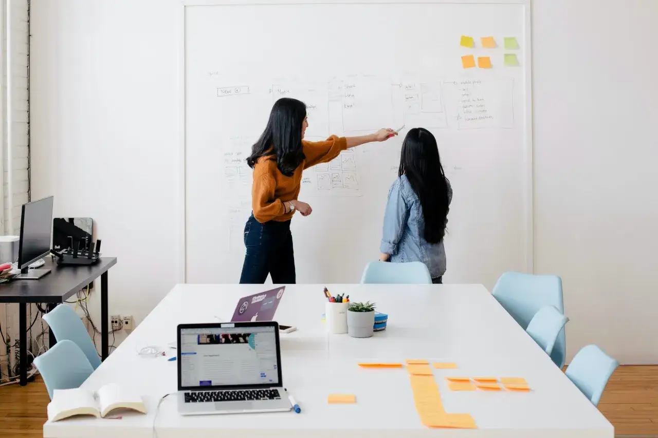 Due donne discutono su una lavagna, pianificando idee per corsi di formazione professionale gratuiti.