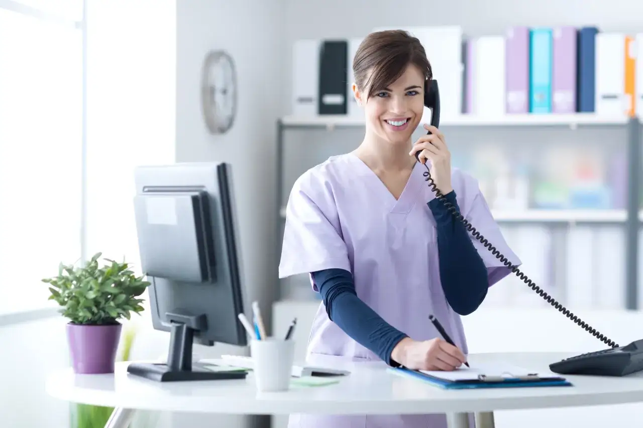 Sorridendo, una segretaria di studio medico al telefono prende appunti. Potrebbe essere un annuncio per offerte lavoro.