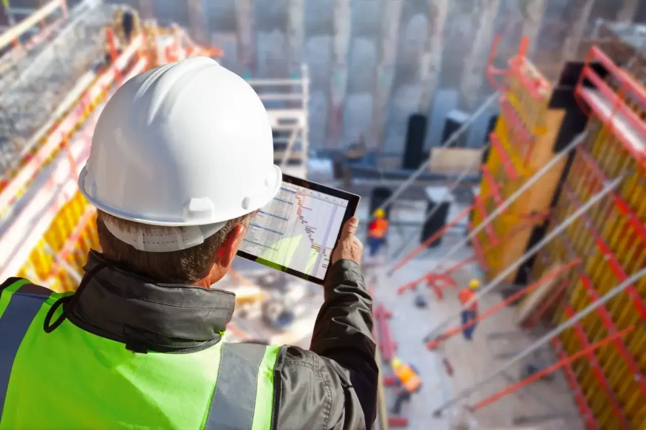 Un site manager con casco bianco e giubbotto ad alta visibilità controlla il progetto su un tablet, mentre operai lavorano in un cantiere edile.