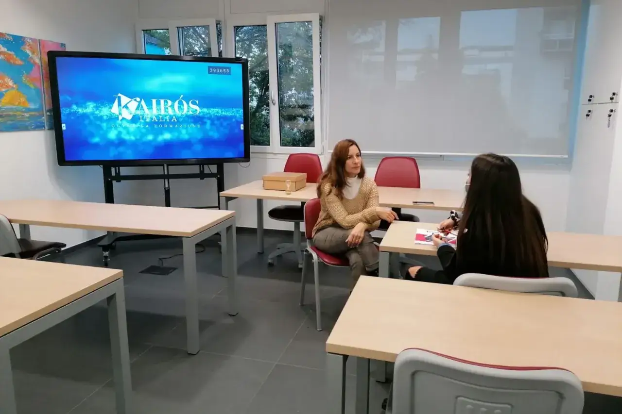 Aula con due studentesse che discutono. Sullo schermo il logo Kairos Italia, che offre corsi gratuiti senza diploma.