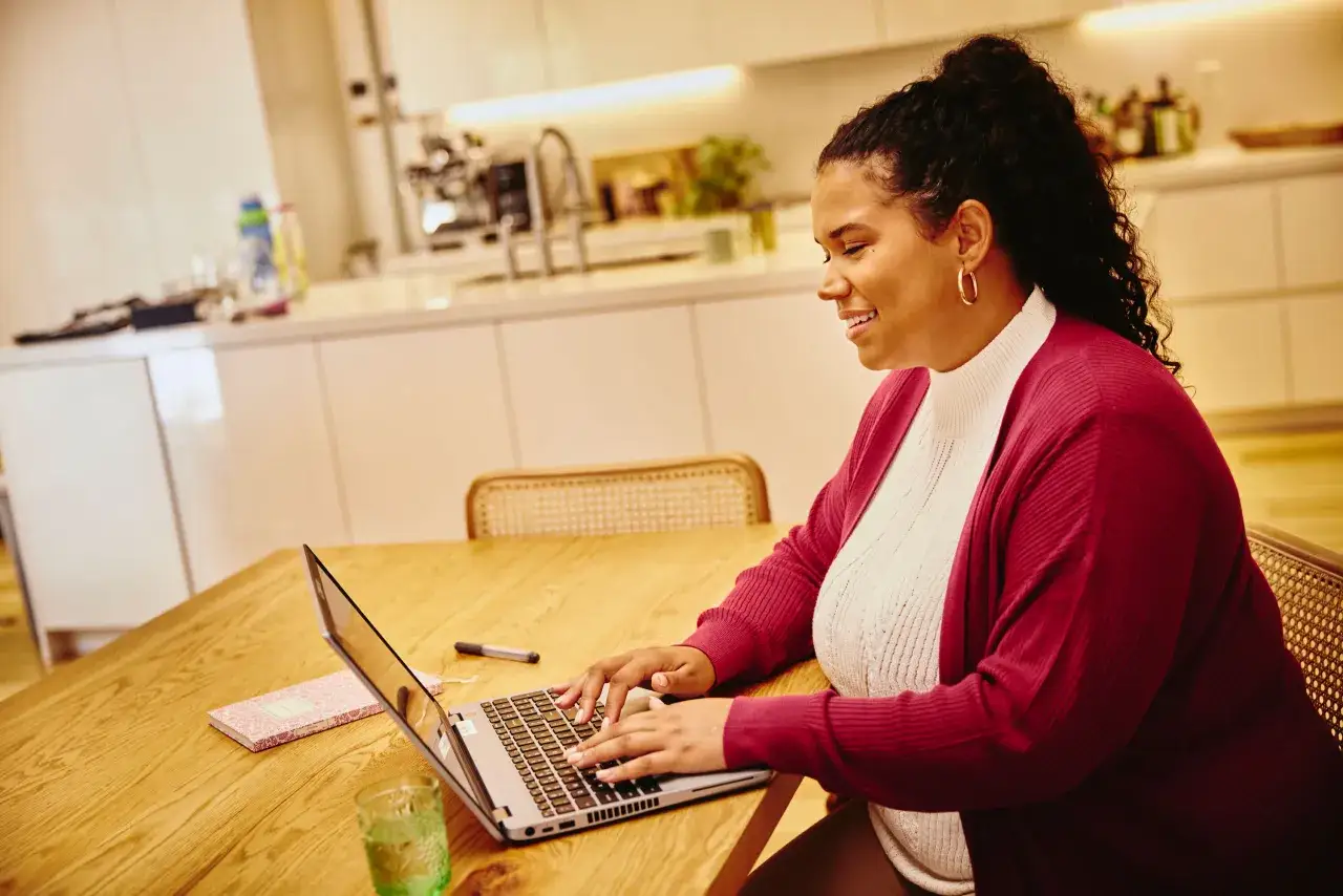 Donna sorridente lavora da casa al suo laptop, con un quaderno e un bicchiere d'acqua sul tavolo di legno.