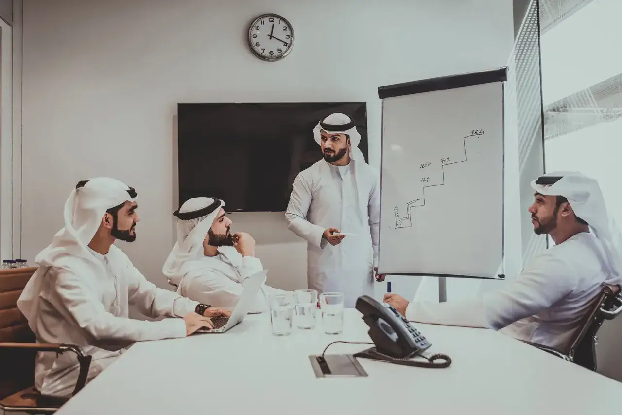 Uomini d'affari in abiti tradizionali discutono strategie di crescita in una sala riunioni, un esempio di come si può lavorare in Qatar.